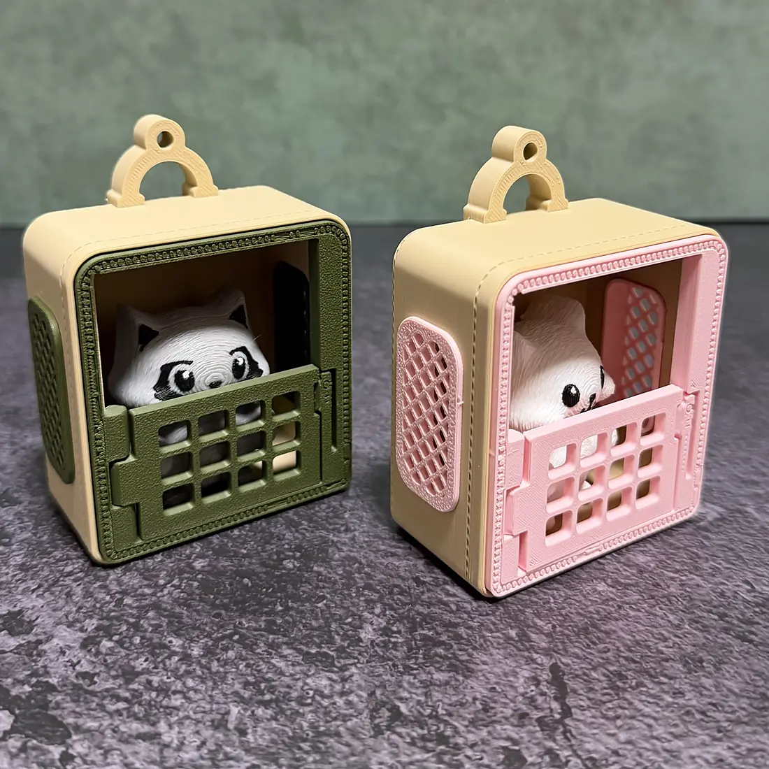 Two printed carrier keychains side by side: a sage one with the Raccoon and a blush one with the Cat peeking over the grate doors, each with lattice side panels and a keychain loop on top